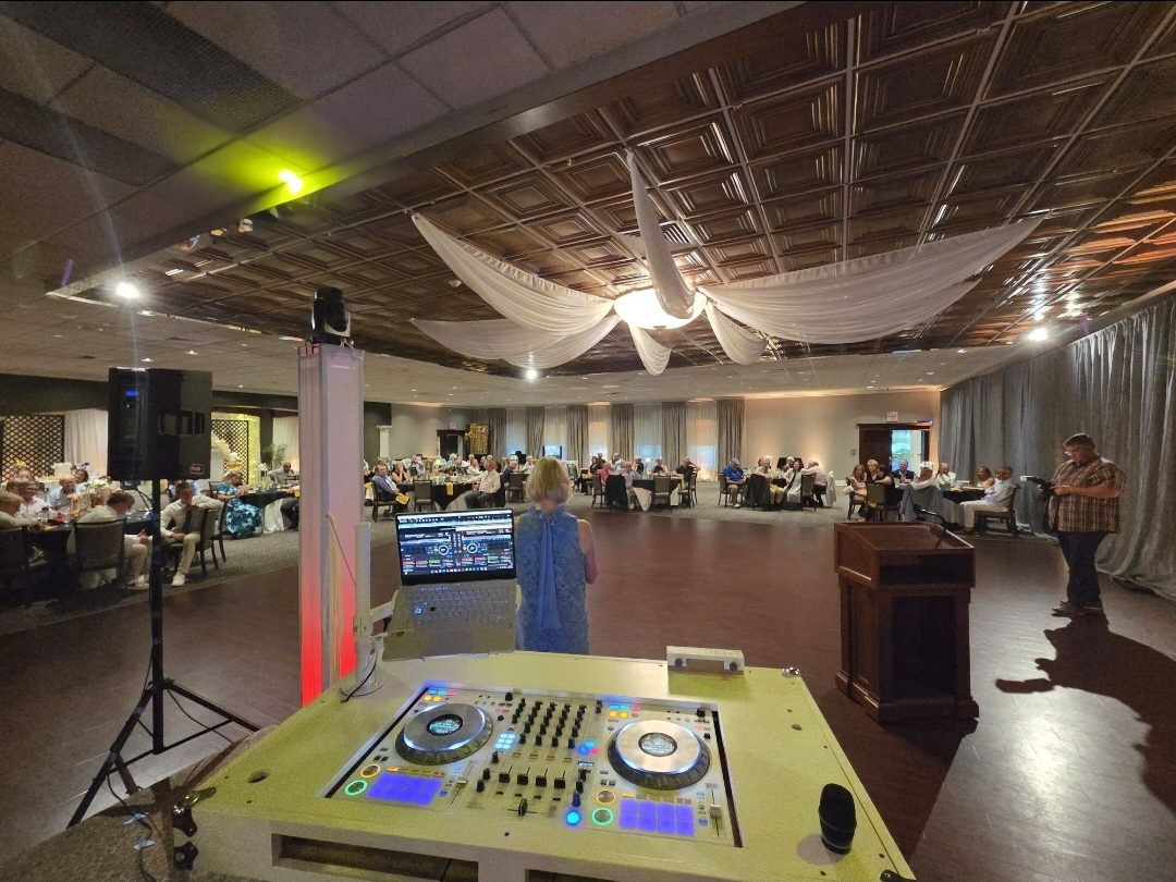 Wide view of guests enjoying an anniversary dinner in a ballroom with draped ceiling, DJ booth, and warm accent lighting