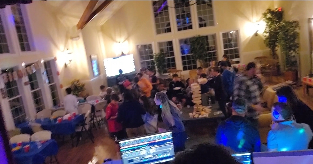 Wide interior shot of a lively birthday party in a bright hall with guests gathered around a giant Jenga game