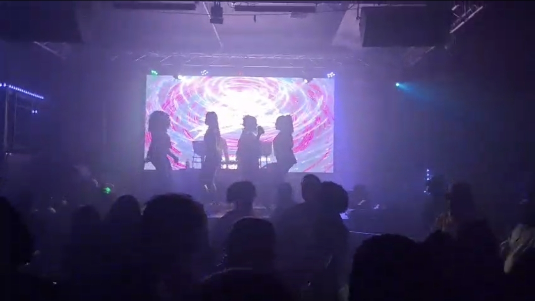 Silhouetted crowd in front of a large LED wall as performers move on stage in a haze-filled nightclub