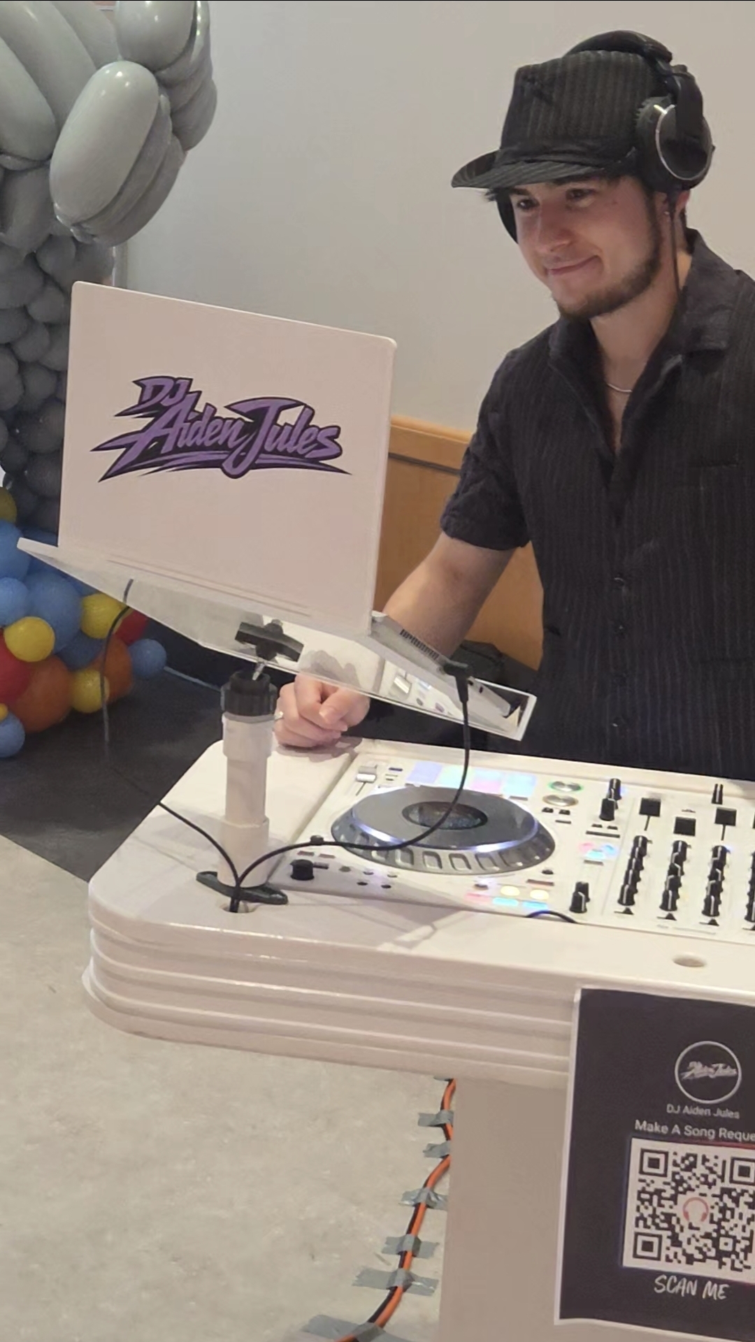 Close-up of DJ Aiden Jules mixing at a college event with a branded booth panel and headphones on