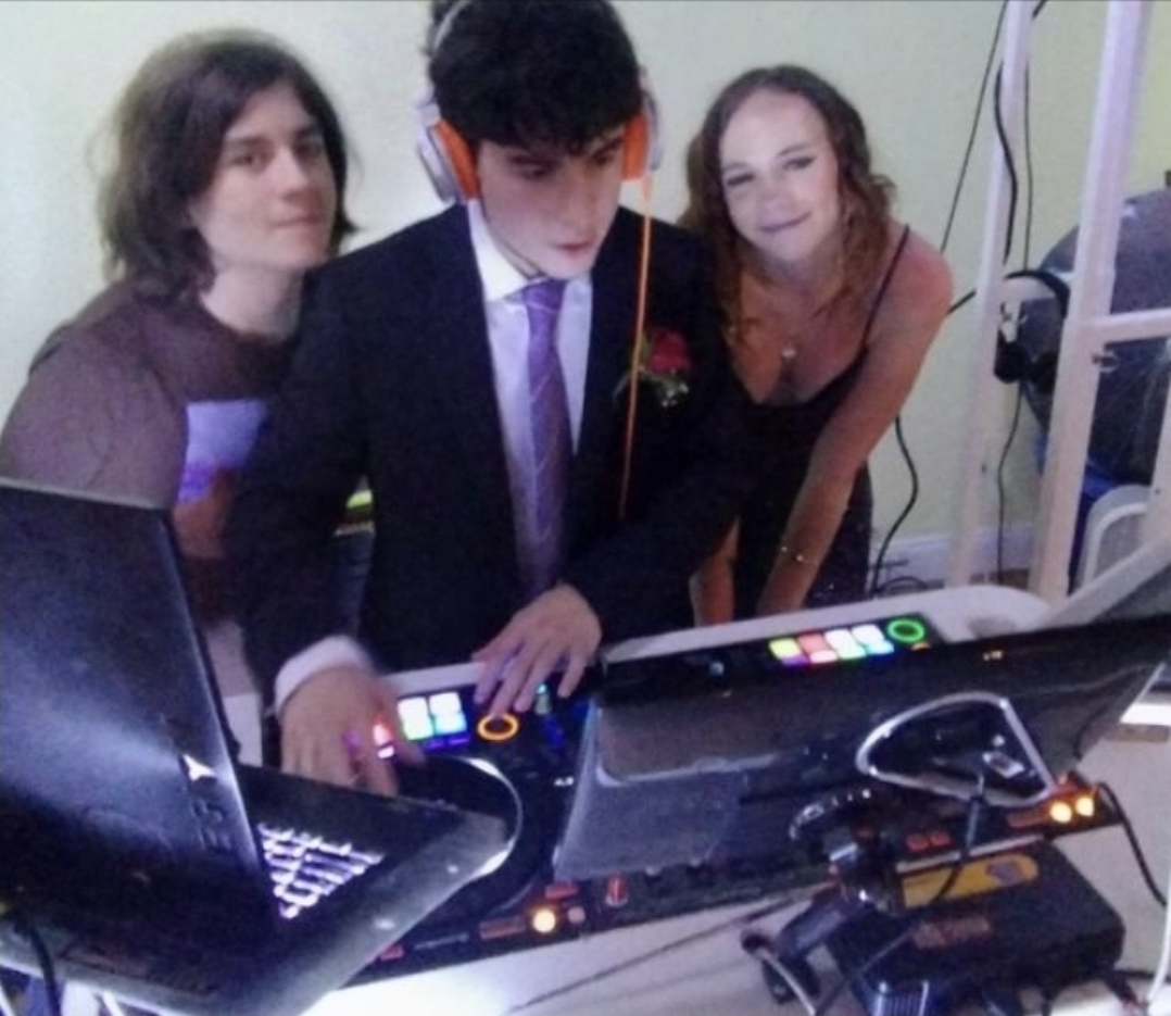 DJ Aiden Jules in a suit behind the controller with two guests smiling beside him at a birthday party