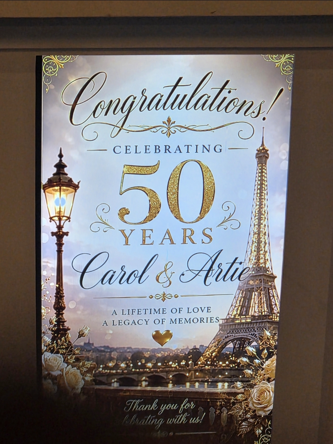 Illuminated 'Congratulations — Celebrating 50 Years, Carol & Artie' Paris-themed anniversary sign with gold lettering and the Eiffel Tower