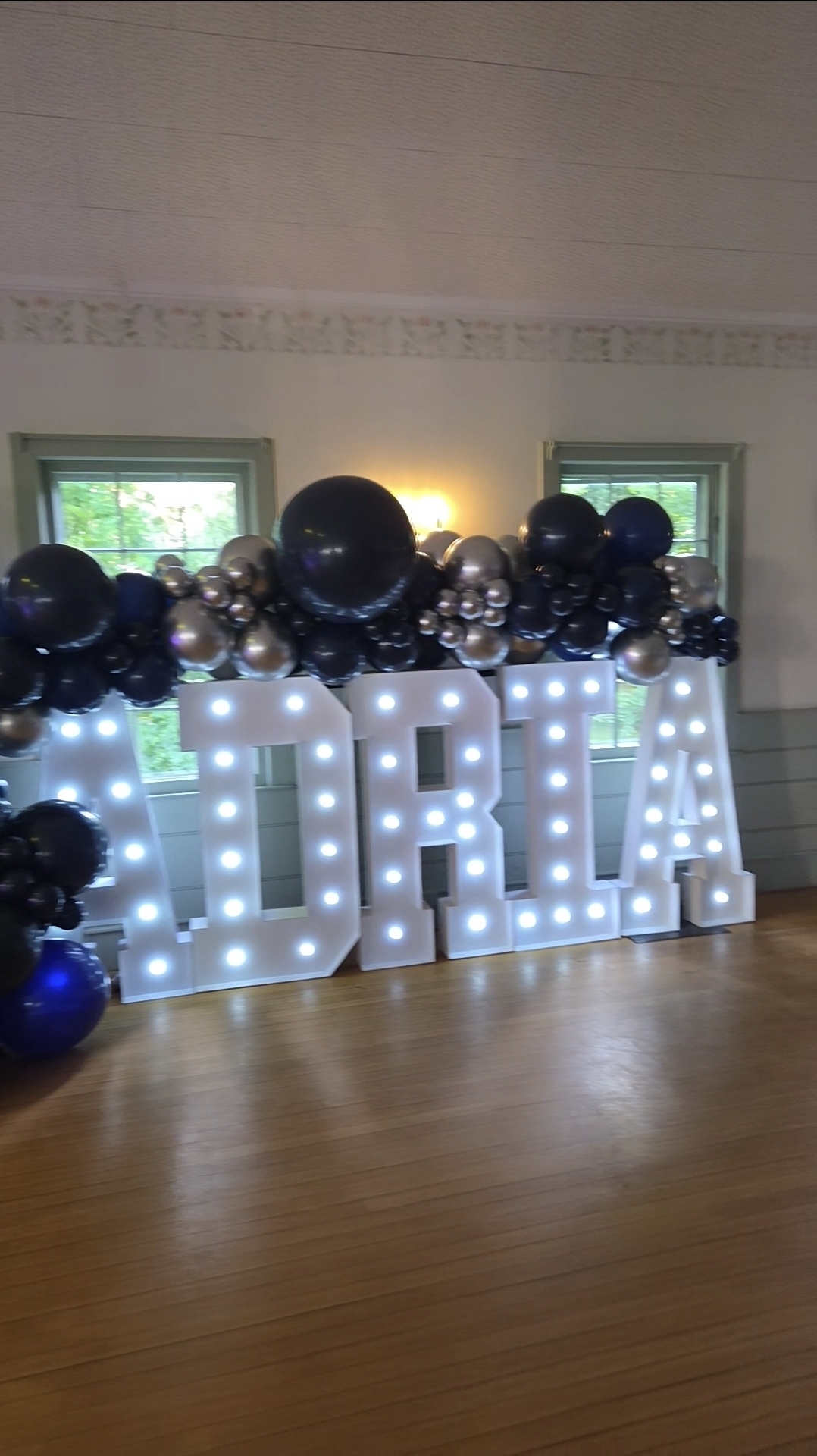 Illuminated white marquee letters spelling ADRIA in front of a black and silver balloon garland